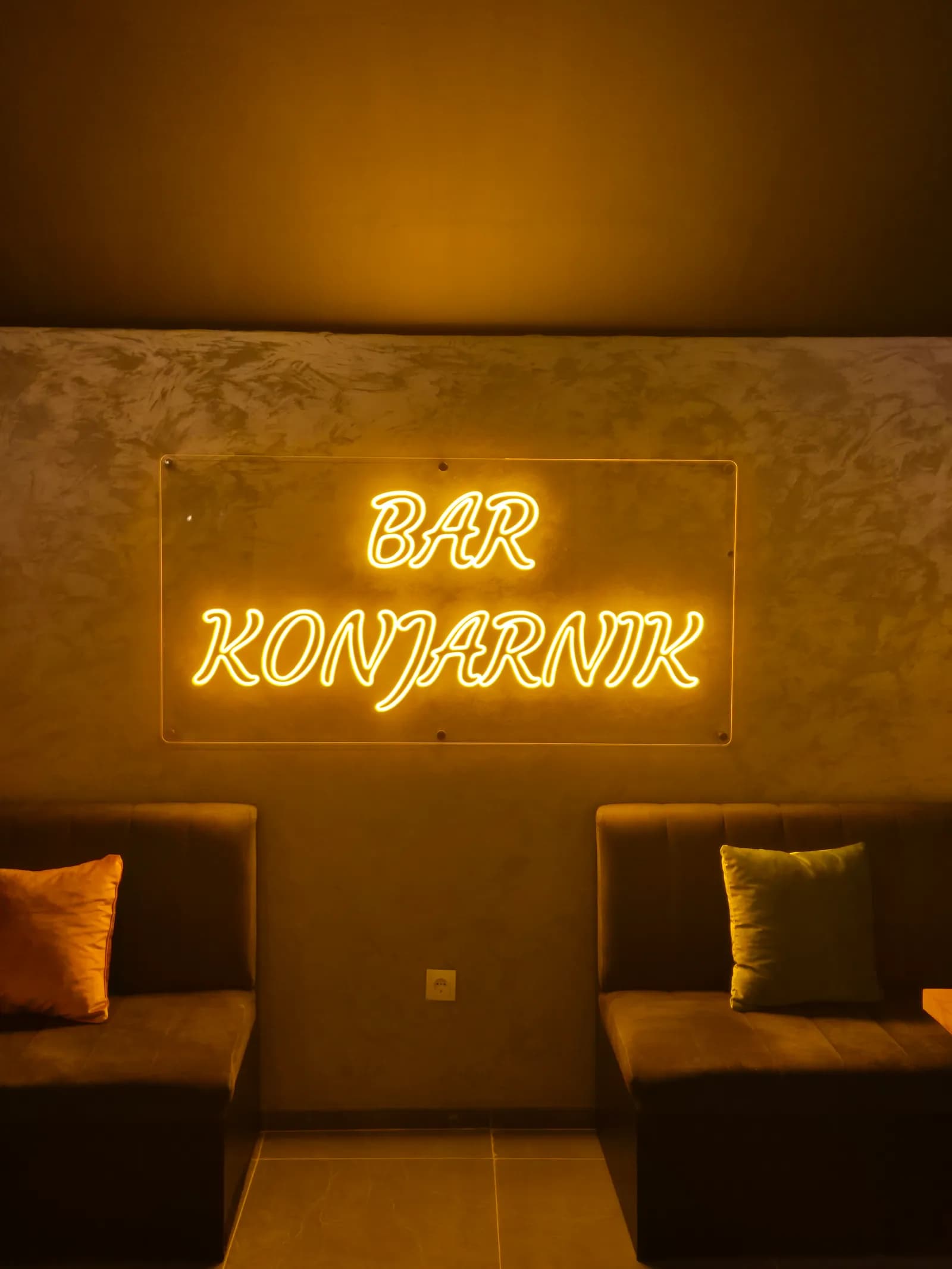 LED neon natpis „Bar Konjarnik" u toploj ćilibarskoj boji, na providnoj kliritnoj ploči iznad sedišta u baru